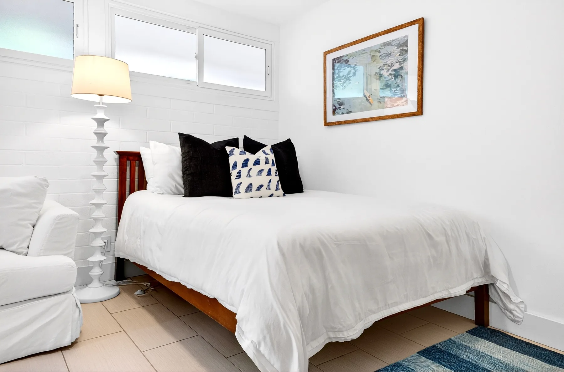 Un dormitorio luminoso con una cama blanca, cojines estampados y un cuadro enmarcado.