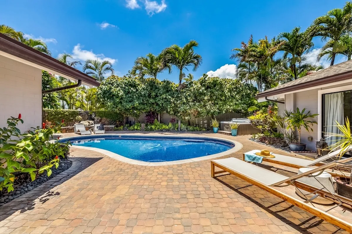 Un patio soleado con piscina, tumbonas, plantas tropicales y palmeras.