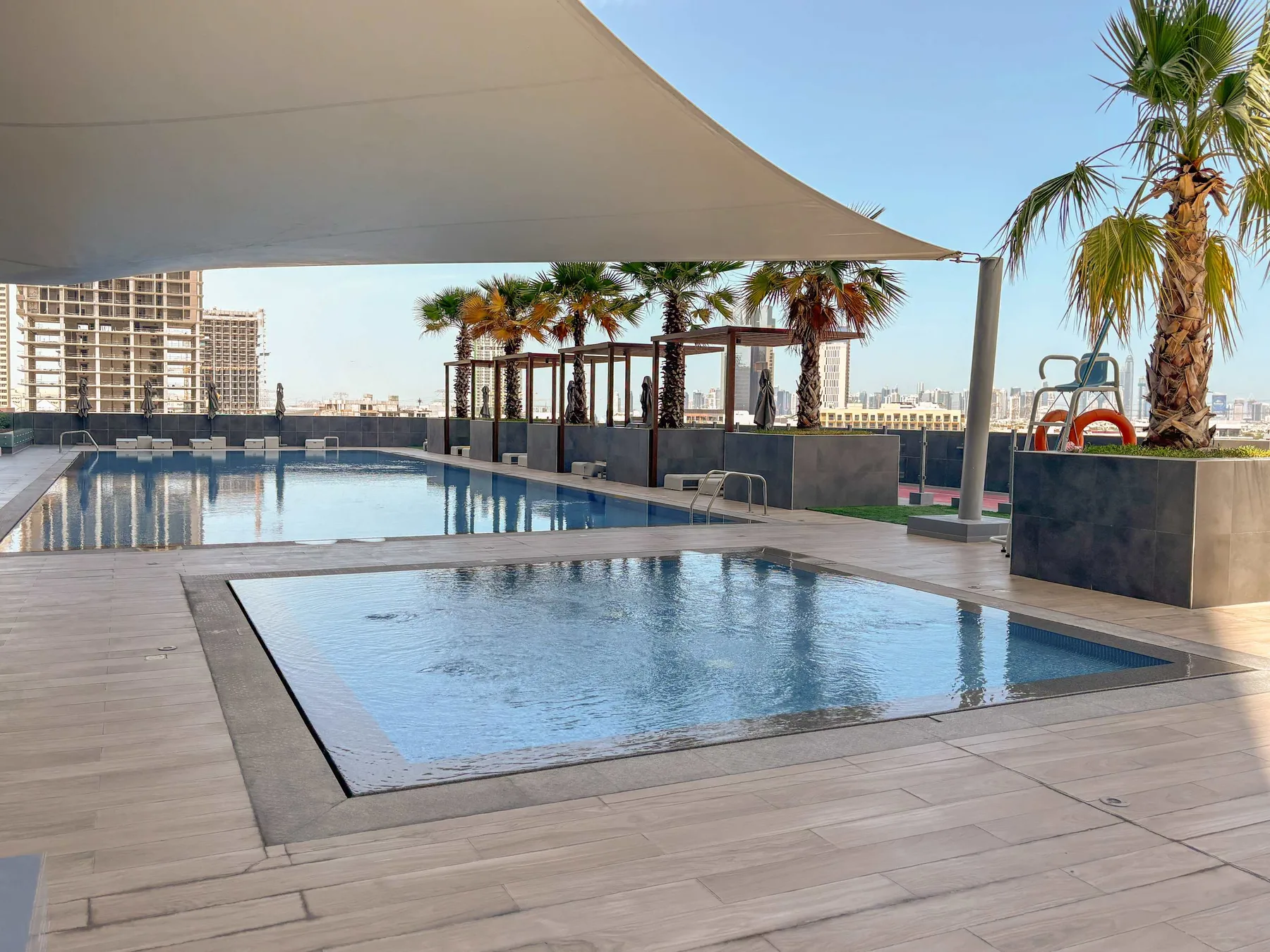 Una zona de piscina al aire libre está bajo una cubierta de sombra, con palmeras cerca.