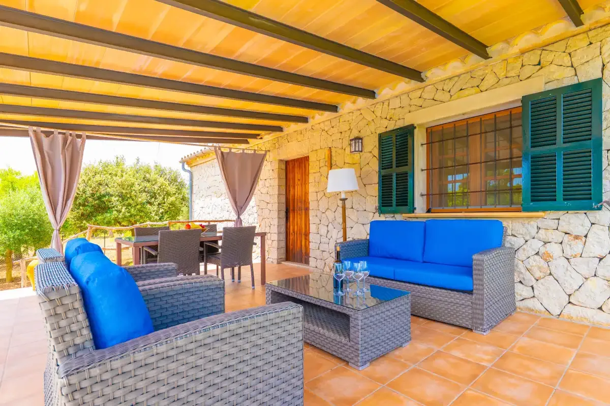 Un patio cubierto tiene asientos de mimbre con cojines azules y una mesa de centro de vidrio.