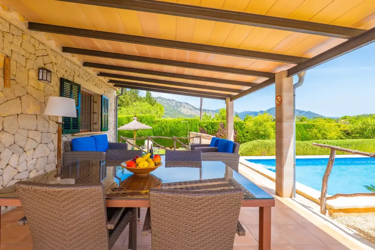Un patio cubierto tiene asientos de mimbre y una mesa junto a una piscina.