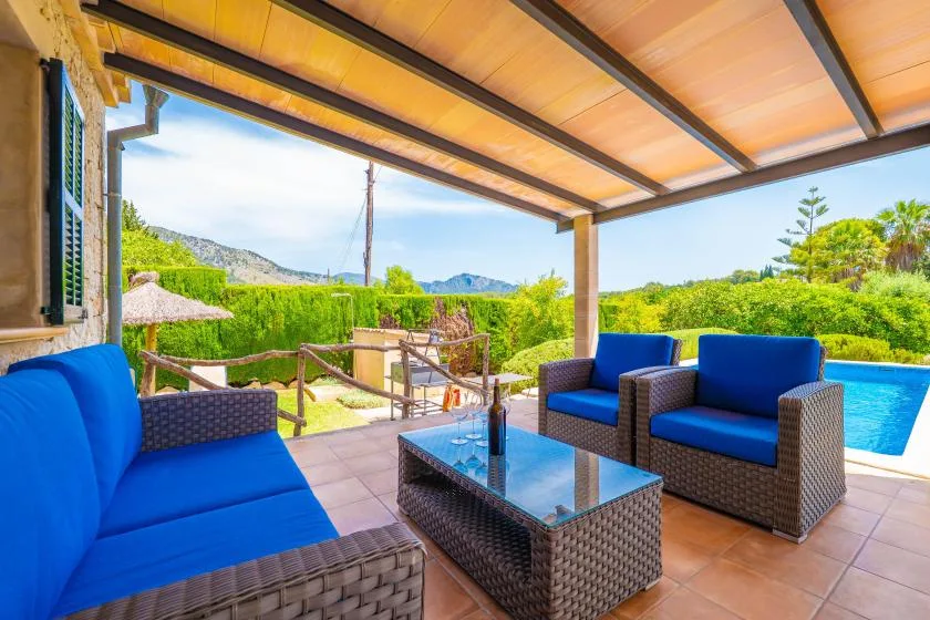 Un patio cubierto tiene asientos azules alrededor de una mesa con tapa de vidrio y una piscina al fondo.