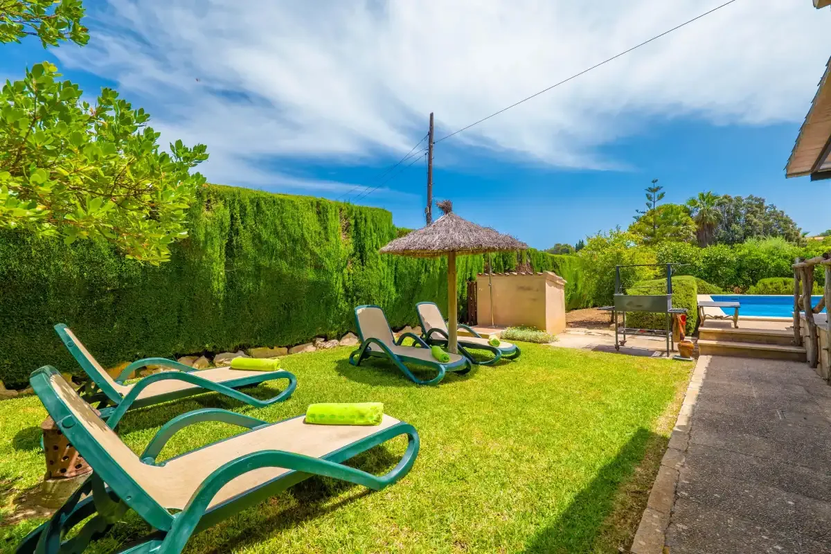 Un patio ajardinado soleado tiene tumbonas verdes, una zona de sombra y una piscina.