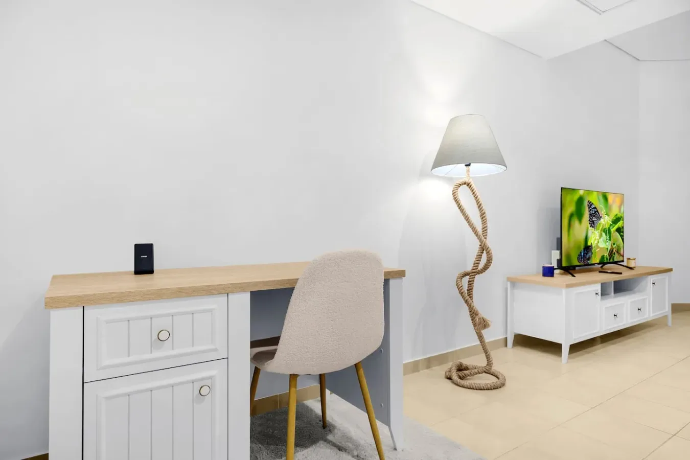 Una habitación luminosa con escritorio, asiento tapizado y televisor sobre un mueble bajo.