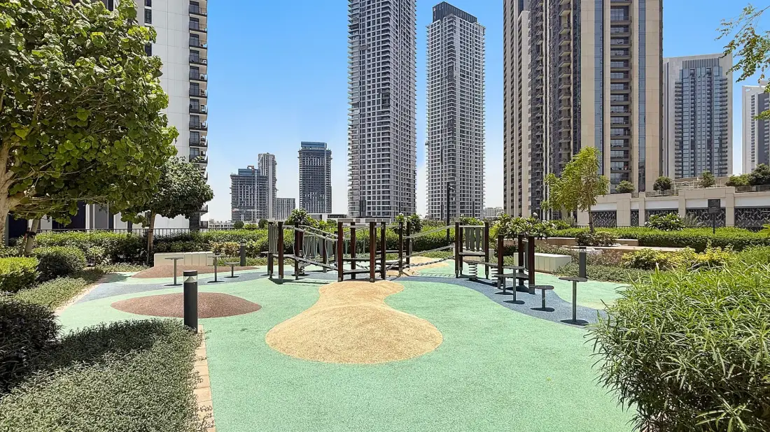 A landscaped outdoor play area with climbing equipment sits among tall residential buildings.