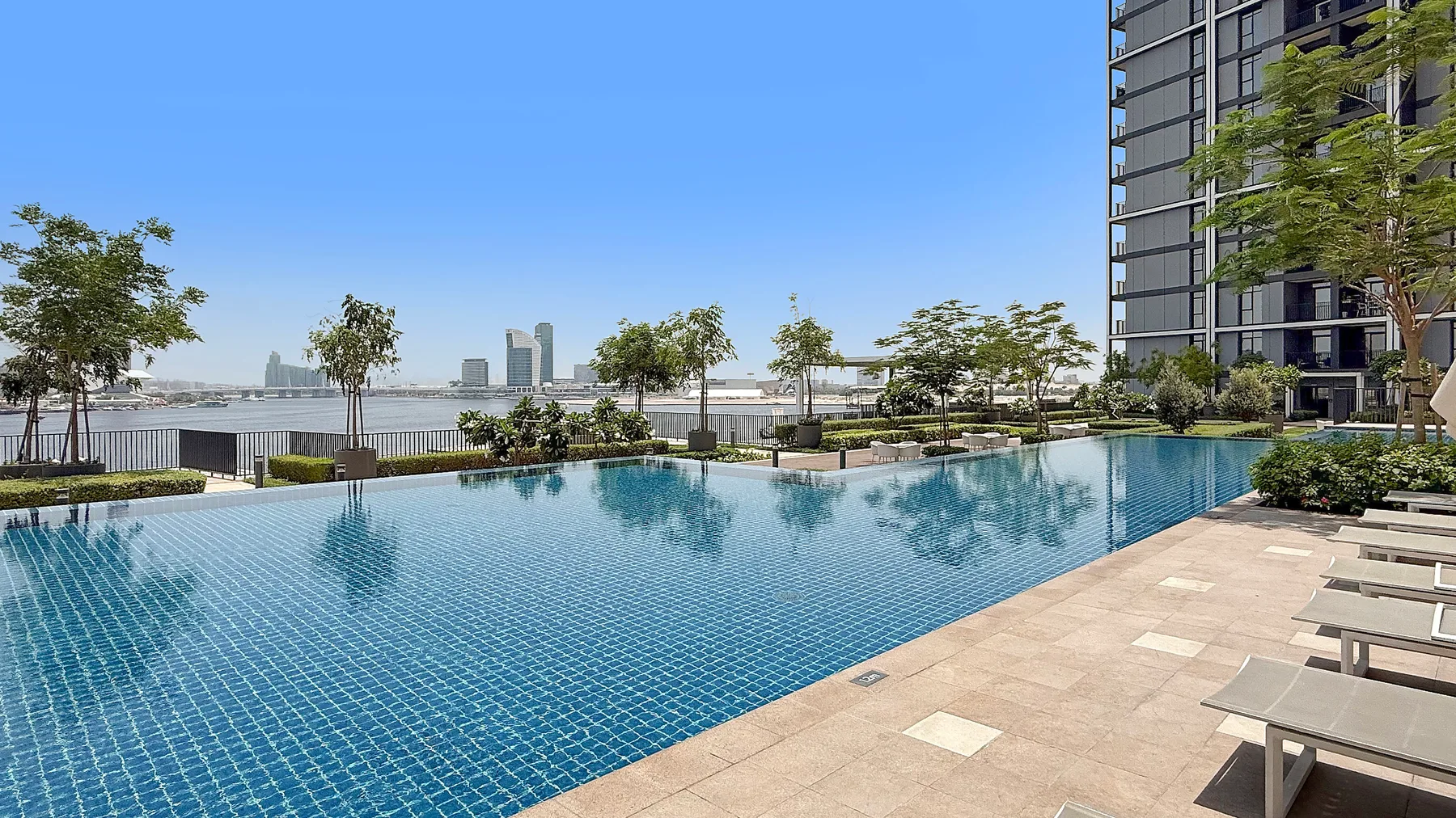 An outdoor swimming pool beside a paved deck, trees, and a high-rise building.