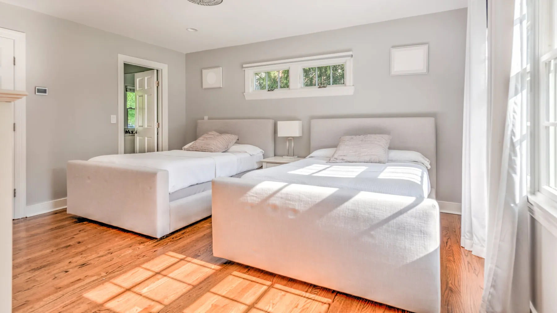 Un dormitorio luminoso con dos camas, paredes claras y luz del sol entrando por las ventanas.
