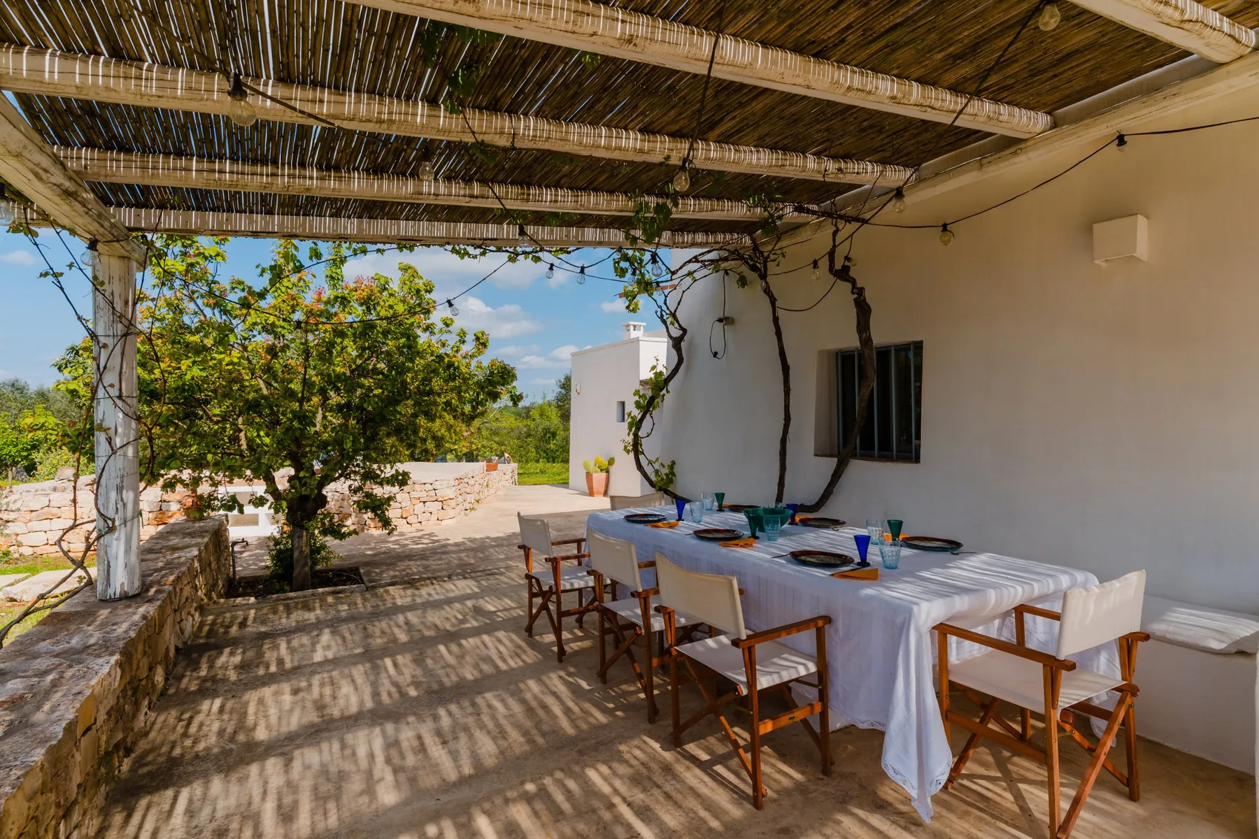 Una zona de comedor exterior a la sombra, con una mesa larga junto a un edificio blanco.