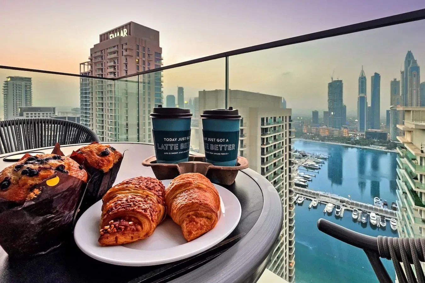 Una mesa en el balcón tiene bollería y café, con vistas a un puerto deportivo y al horizonte urbano.