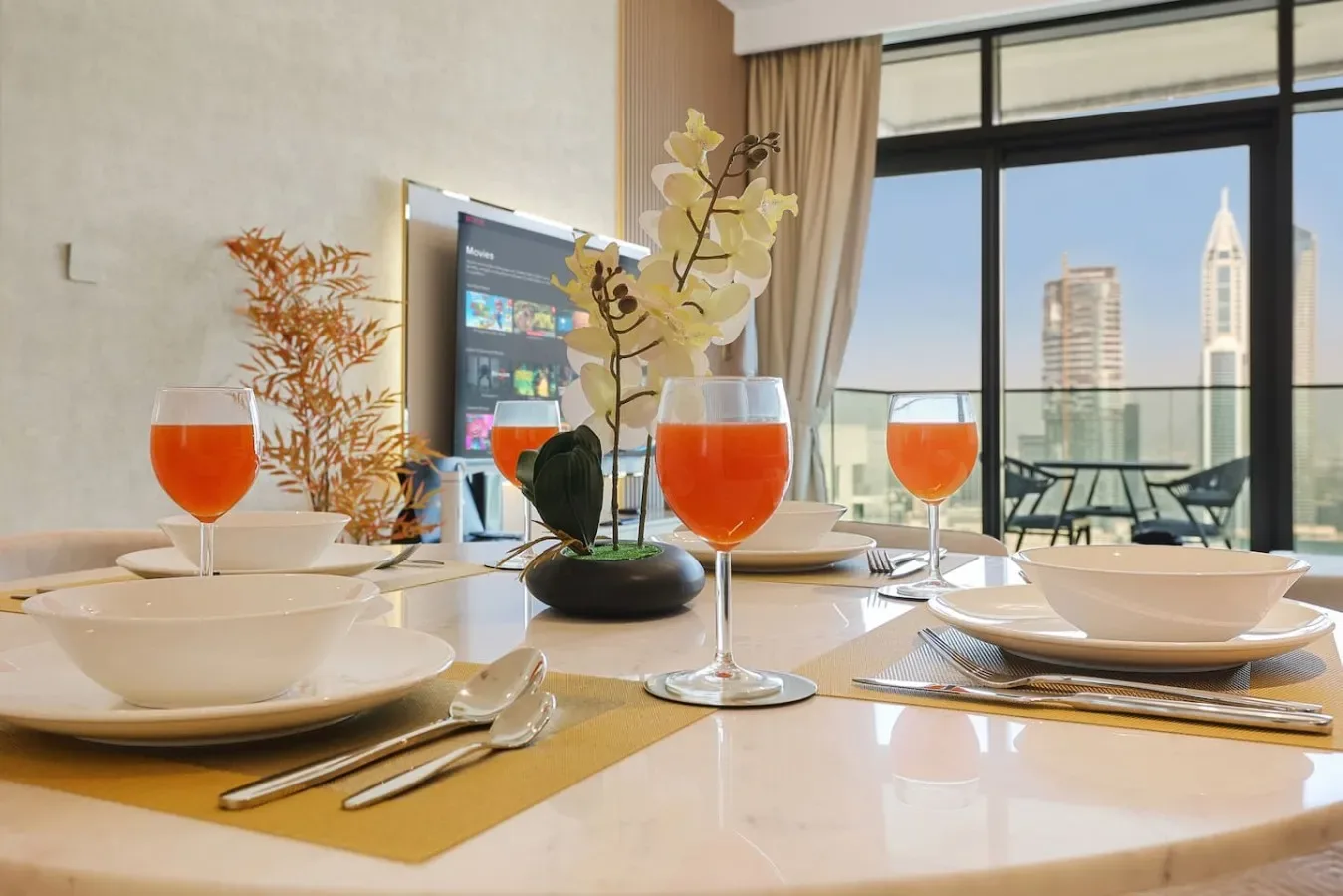 Una mesa está puesta con platos, bebidas naranjas y flores junto a una ventana grande.