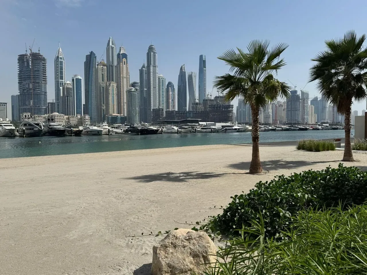 Un paseo costero de arena con palmeras, embarcaciones y un horizonte urbano moderno al otro lado del agua.