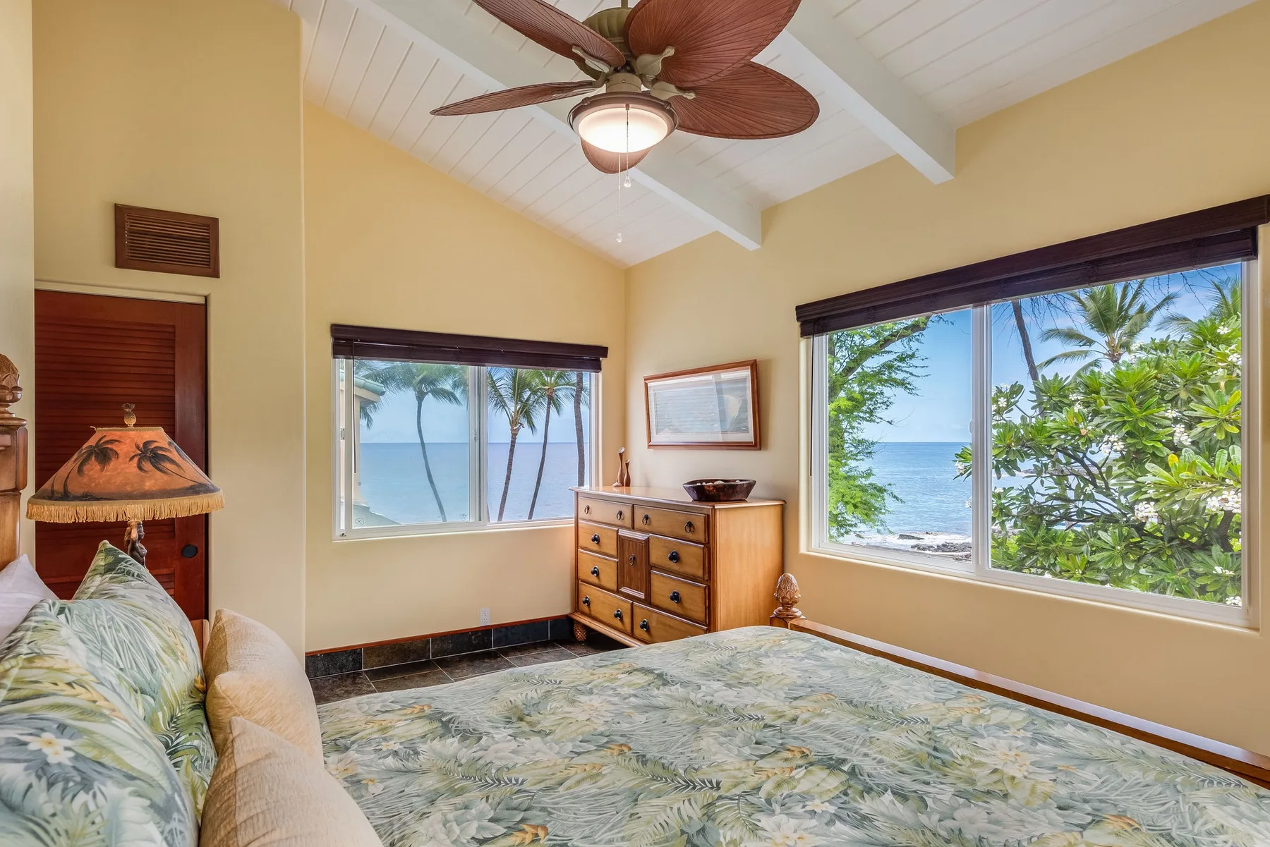 Un dormitorio con cama y cómoda junto a ventanas con vistas a vegetación tropical y al mar.