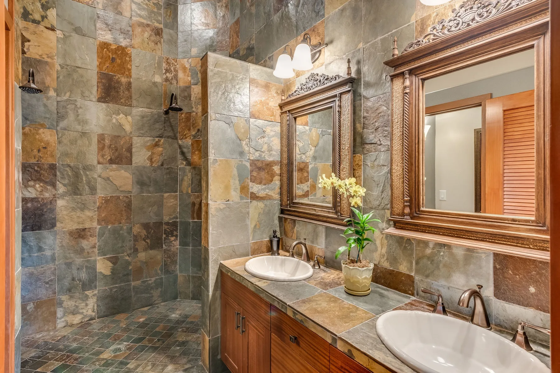 Un baño alicatado con lavabo doble, espejos y flores en una maceta.