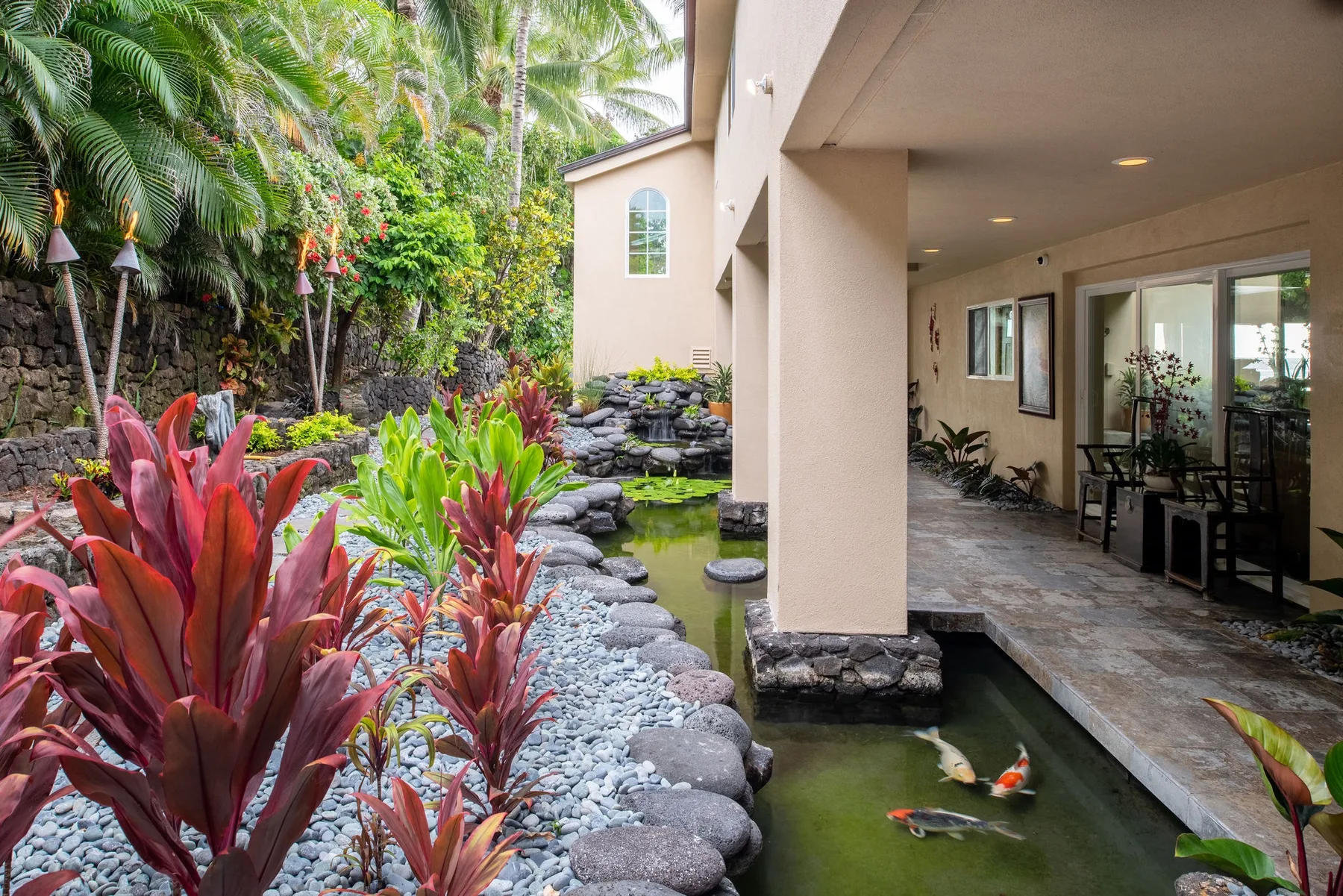Un patio ajardinado con estanque, bordes de piedra y plantas tropicales junto a un pasillo cubierto.