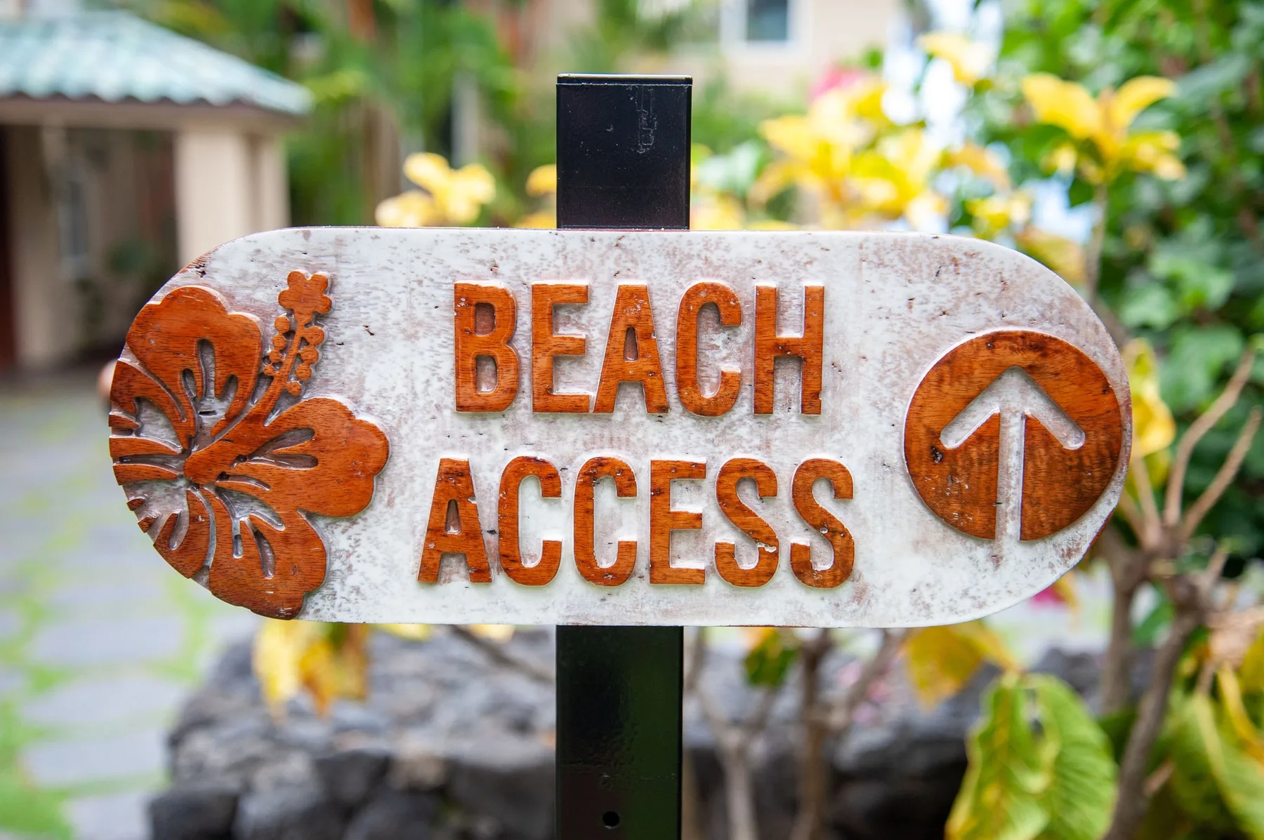 Un letrero de jardín señala el acceso a la playa, rodeado de flores y vegetación tropical.