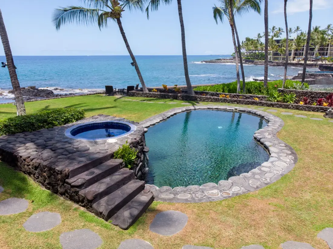 Una piscina de forma irregular y un spa circular están en un césped junto al océano.