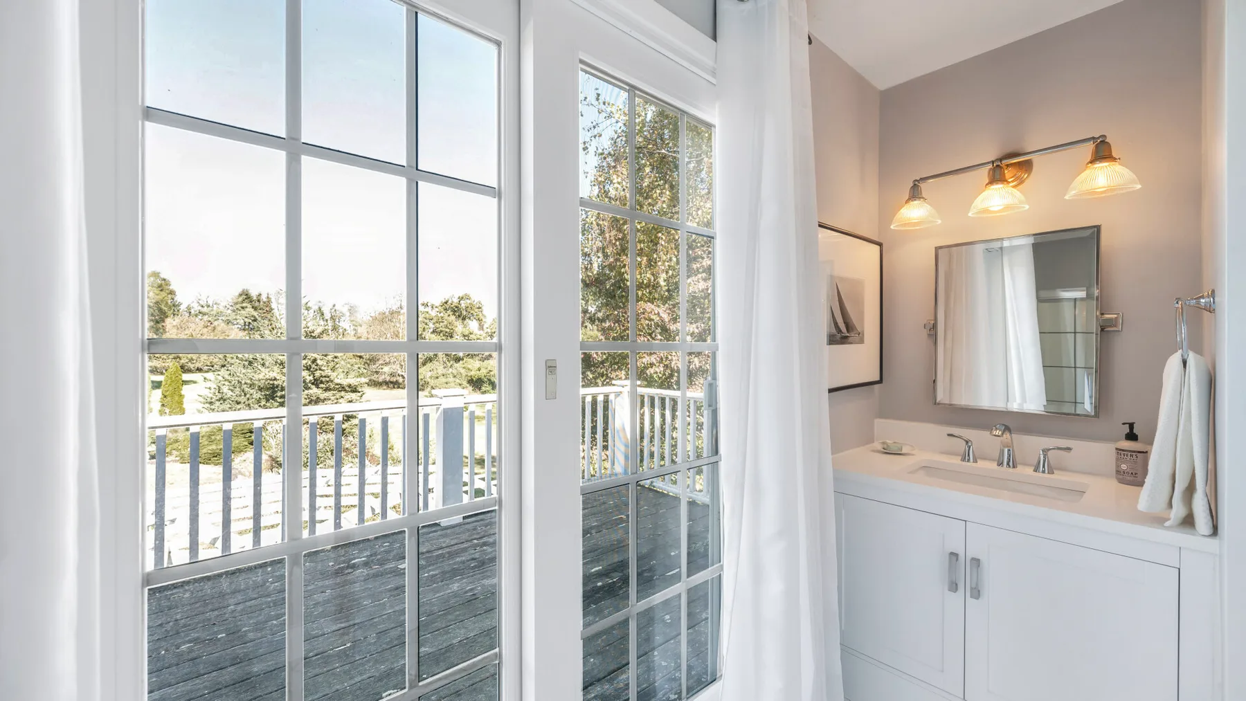 A bright bathroom vanity with a wide mirror beside tall windows overlooking a balcony.