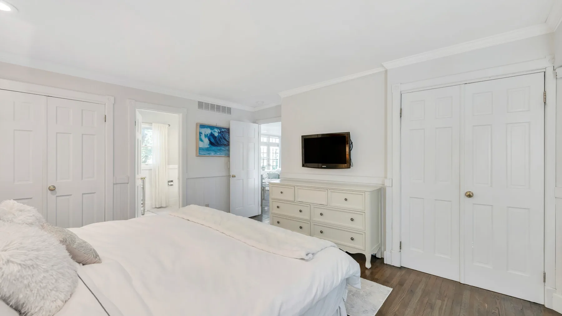 A bright bedroom with a neatly made bed and a dresser beneath a wall-mounted television.