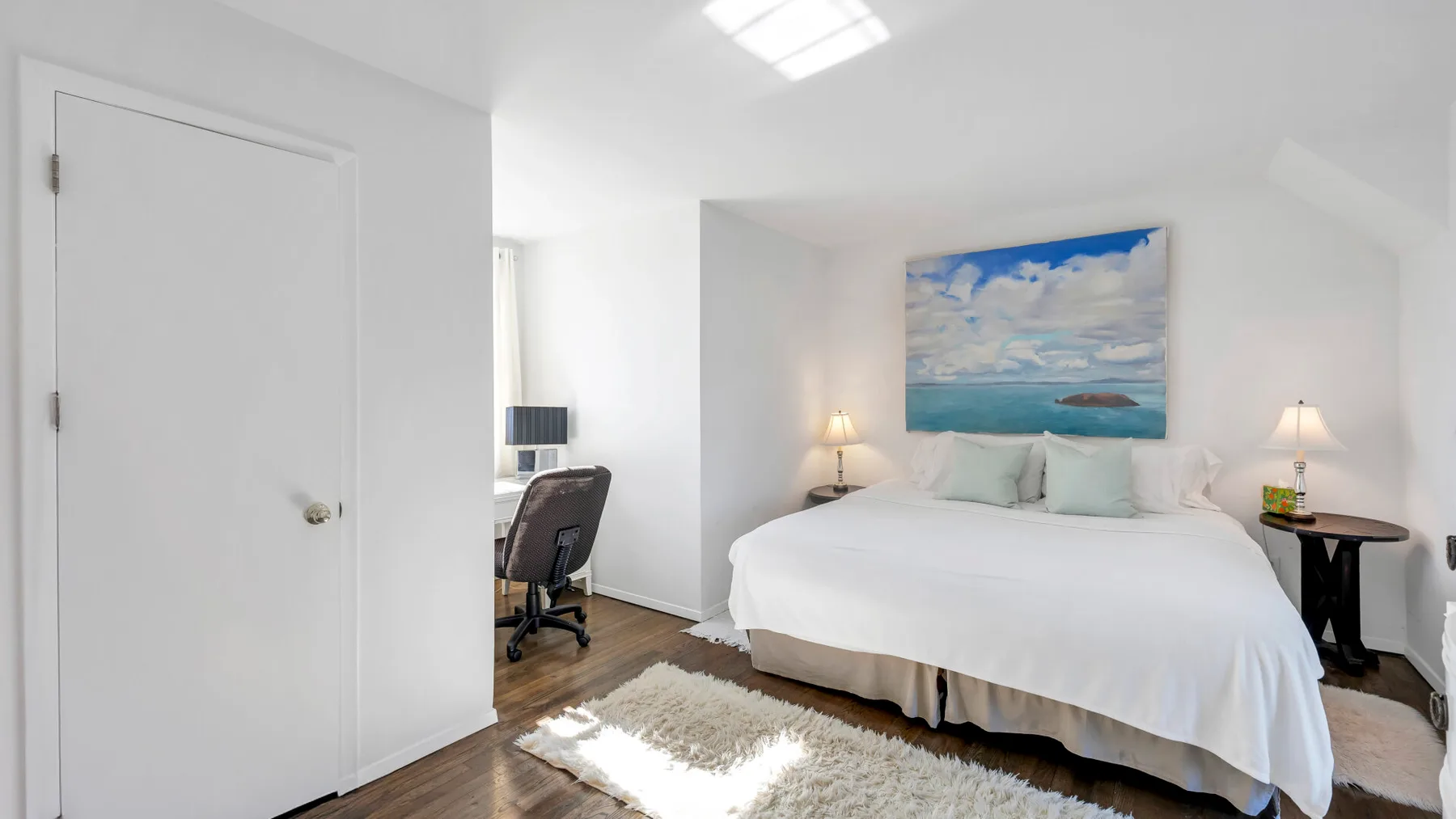 A bright bedroom with a white bed and a coastal painting above it.