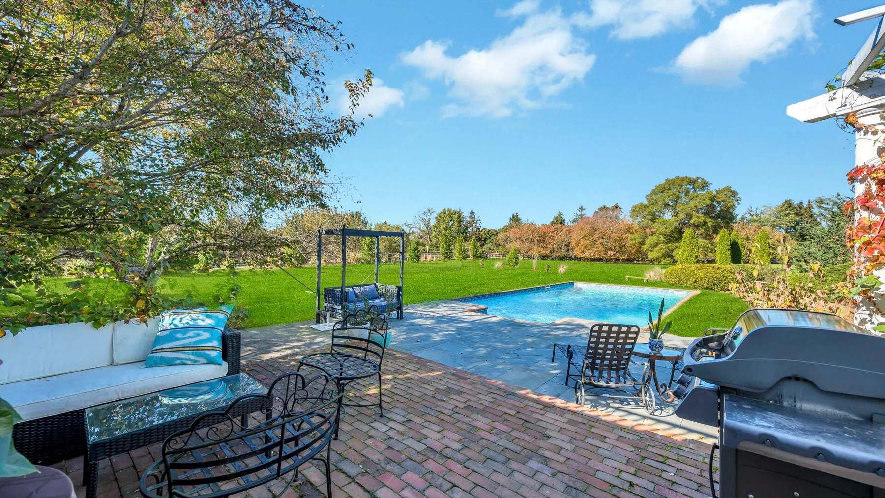 Piscina al aire libre junto a un patio de ladrillo, con asientos y vegetación.