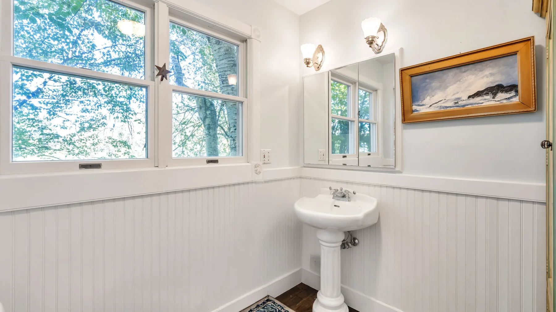 A bright bathroom with a pedestal sink, large windows, and framed artwork.