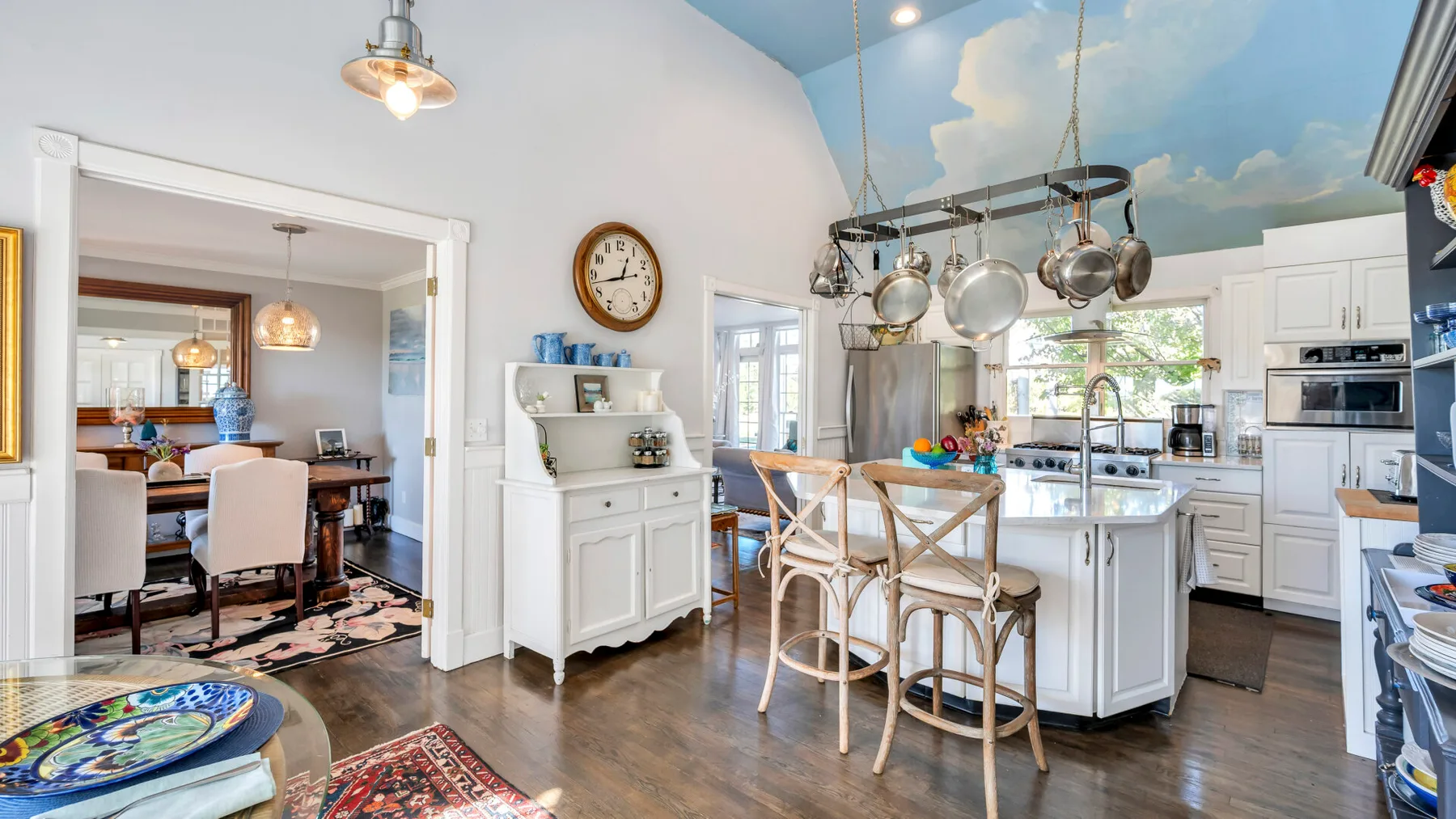 A bright kitchen with a central island and hanging cookware beneath a painted sky ceiling.