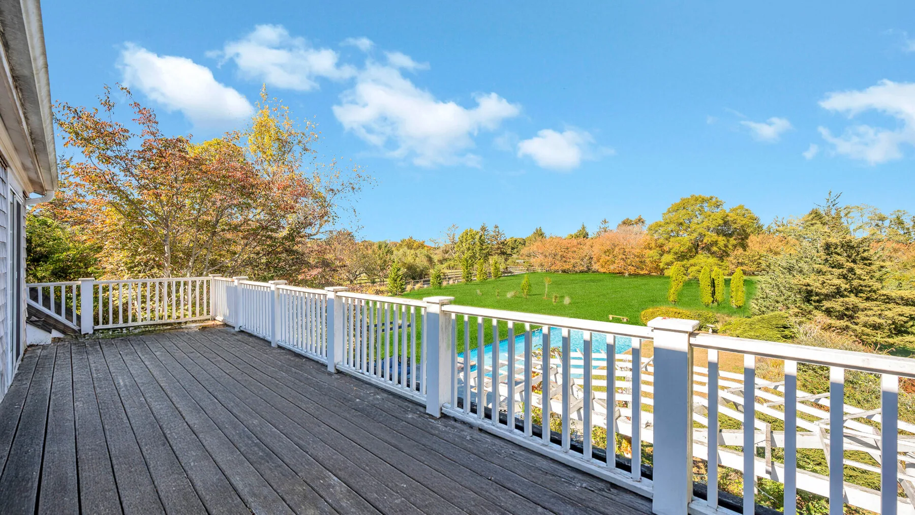 A wooden deck overlooks a green lawn and trees under a blue sky.