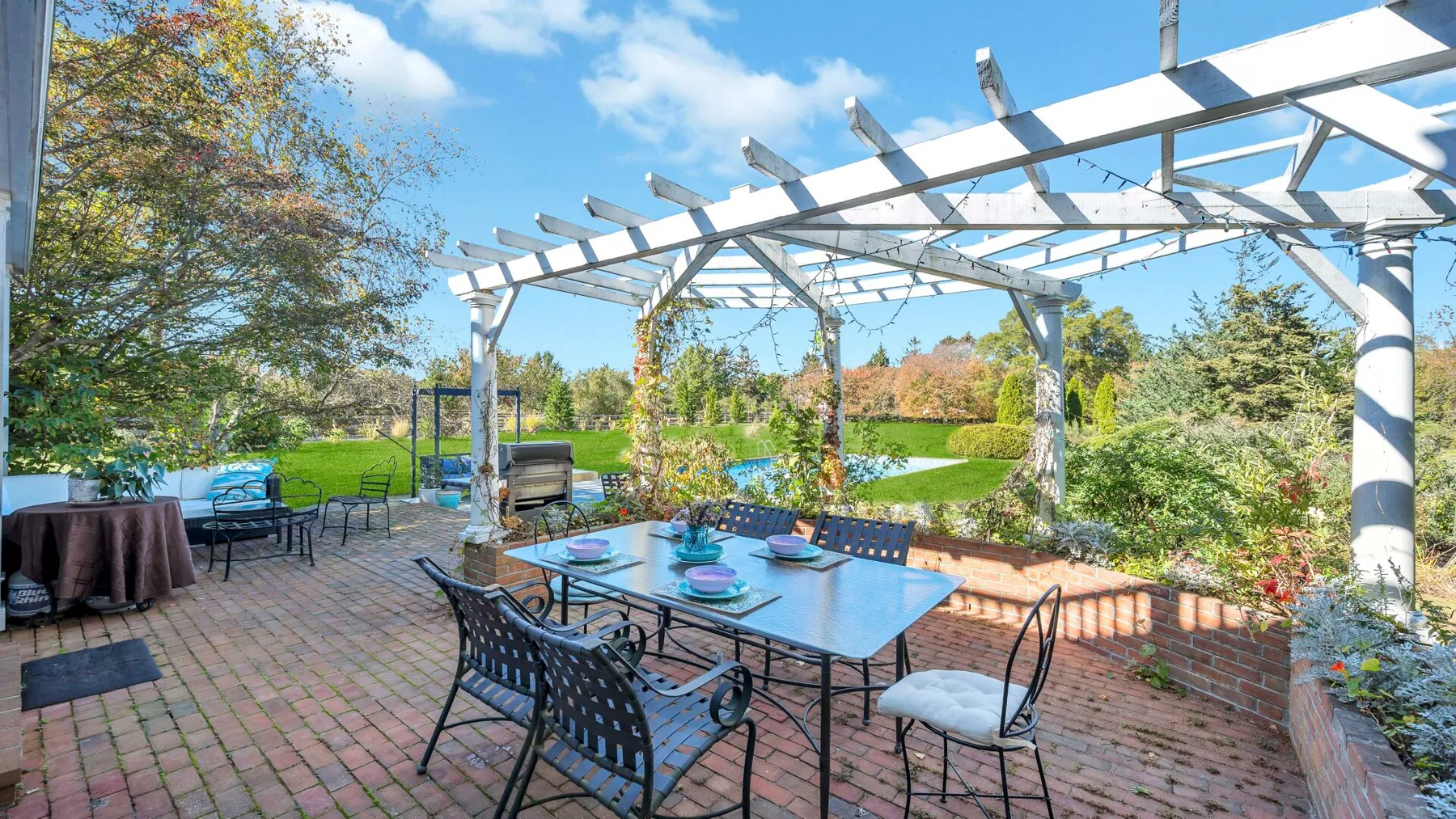 A brick patio with outdoor dining furniture sits beneath a white pergola, overlooking a garden.