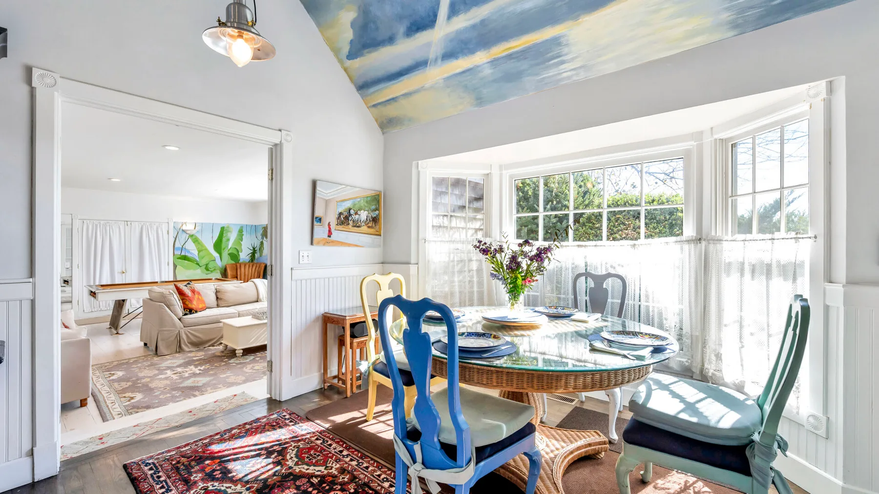 A bright dining area with a round table beside large windows and a colorful painted ceiling.