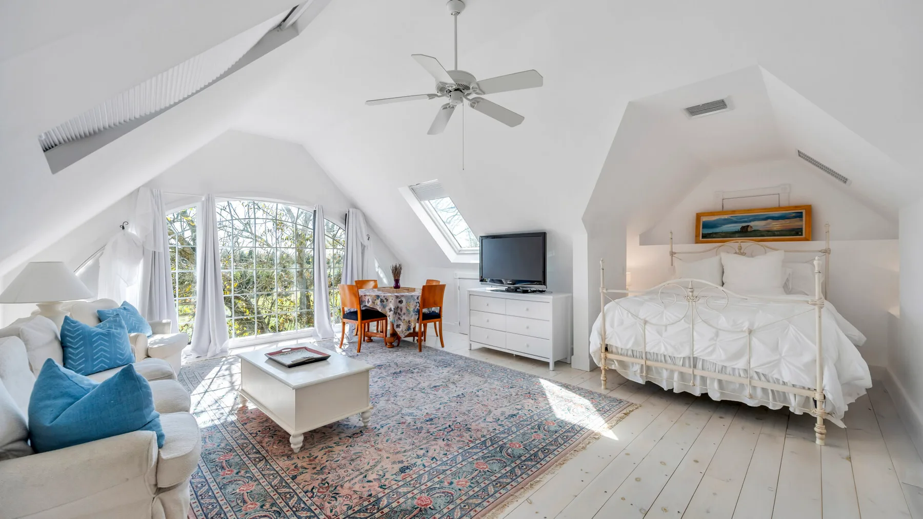 A bright attic bedroom has a bed, seating, and a dining area beneath a sloped ceiling.