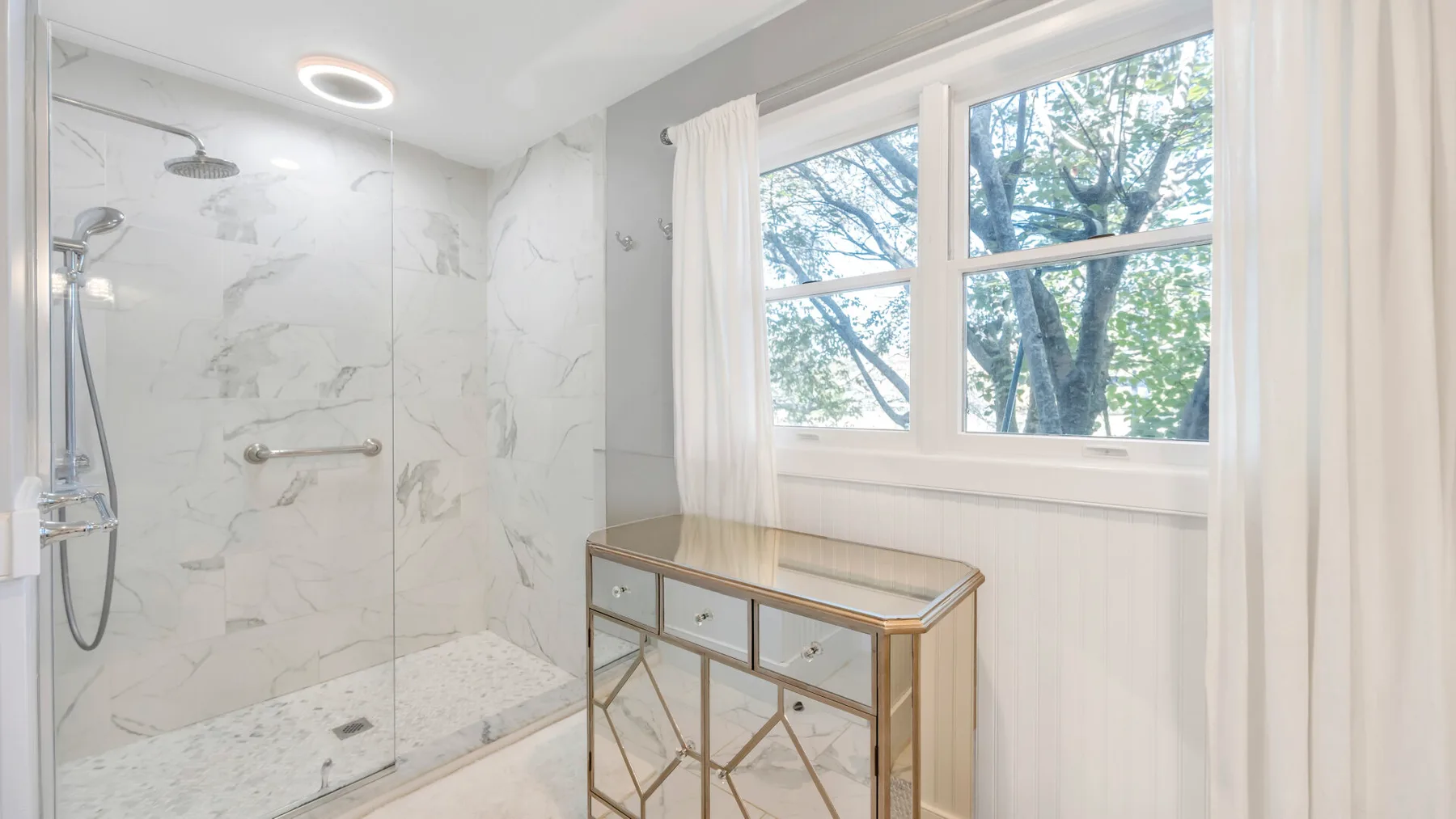 A bright bathroom has a glass shower, a window, and a small vanity.