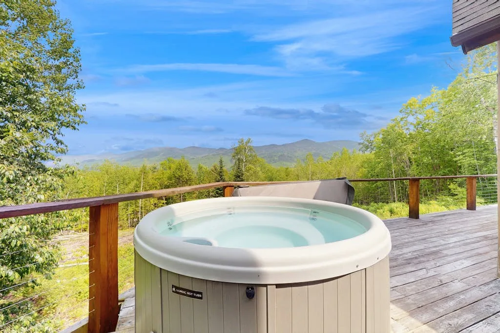A hot tub sits on a wooden deck overlooking a green, mountainous landscape.