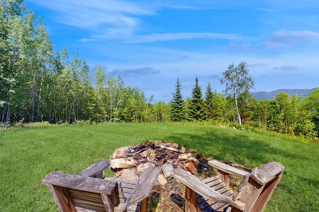 An outdoor seating area surrounds a stone fire pit in a grassy clearing.