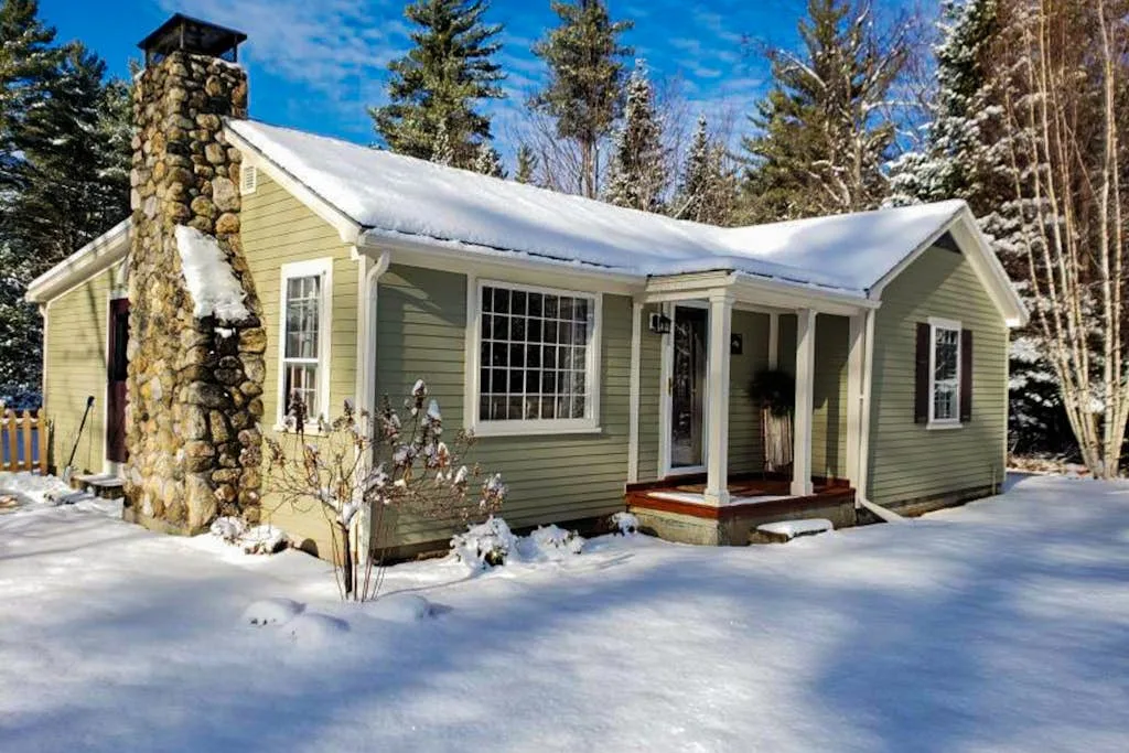 A snow-covered house with a stone chimney stands among trees.
