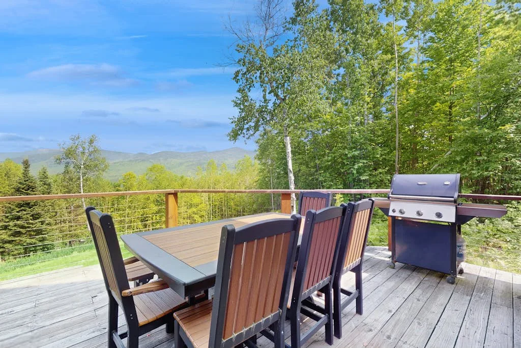 An outdoor dining table and grill sit on a wooden deck overlooking a tree-filled landscape.