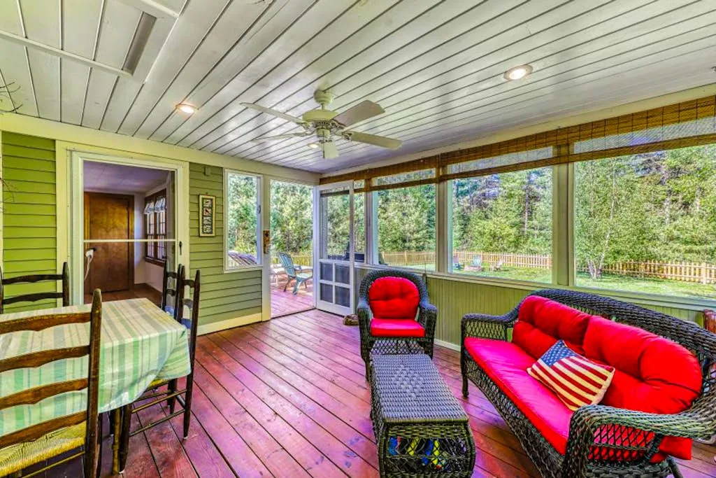 A bright enclosed porch has wicker seating with red cushions and windows overlooking trees.