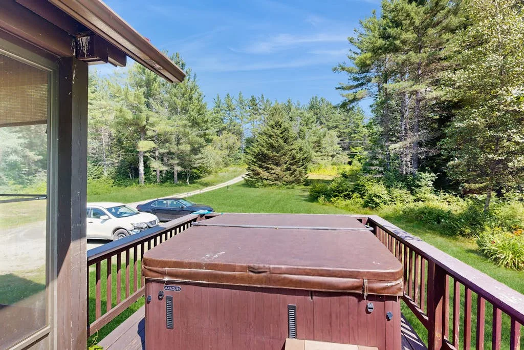 An outdoor deck with a covered hot tub overlooking a green, tree-lined landscape.