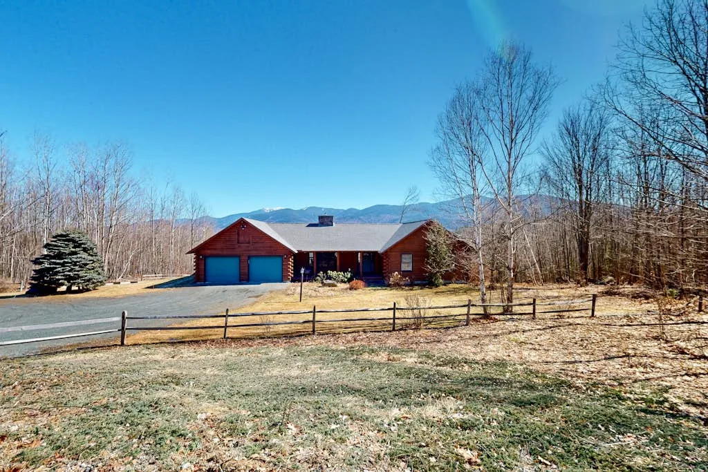 A red house with a blue garage stands beside a fenced yard, with mountains behind it.