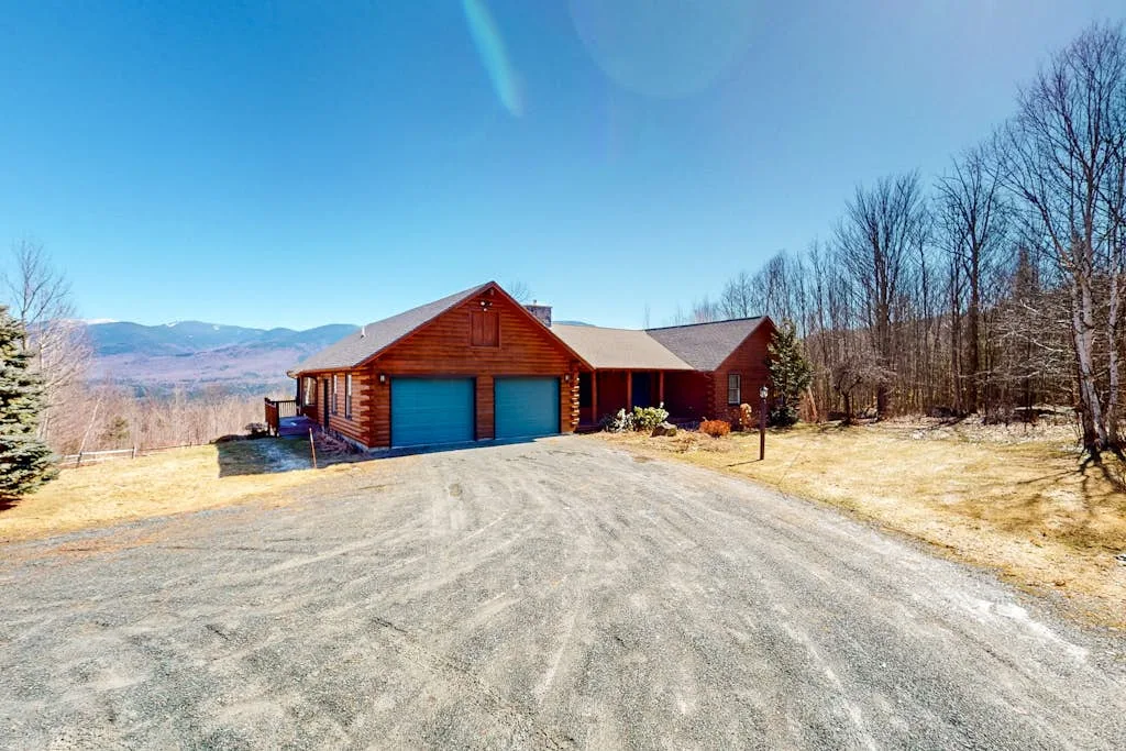 A wooden house with a driveway sits beside leafless trees and open mountain views.