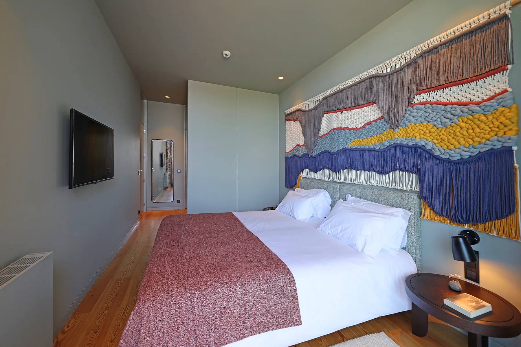 A bedroom with white bedding and a colorful woven wall hanging above the bed.