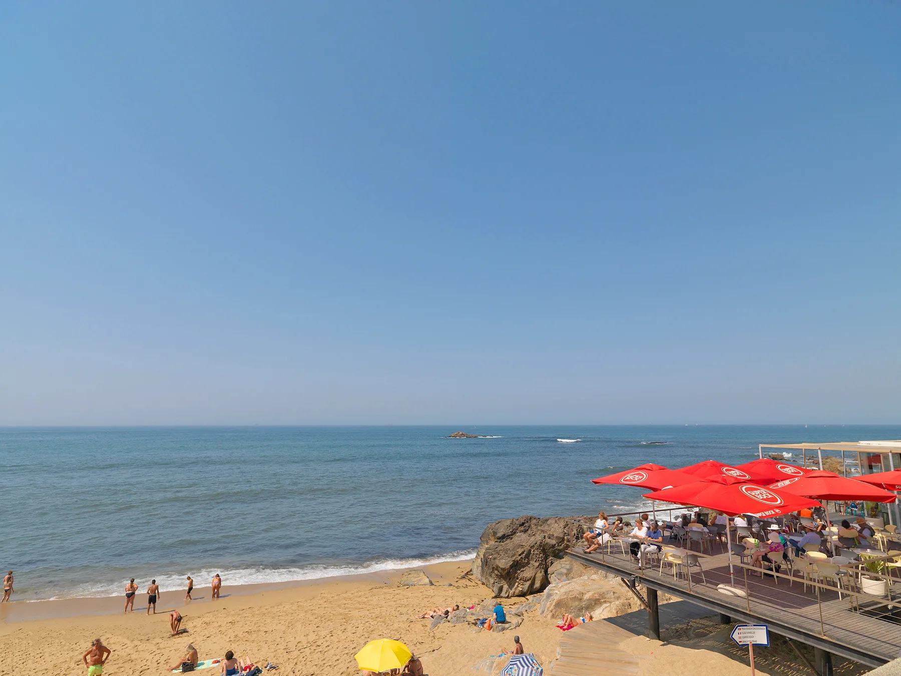 A sandy beach meets calm water beside a seaside terrace with red umbrellas.