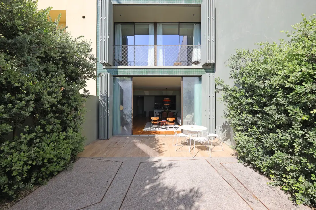 A modern building entrance opens onto a paved patio with outdoor seating.
