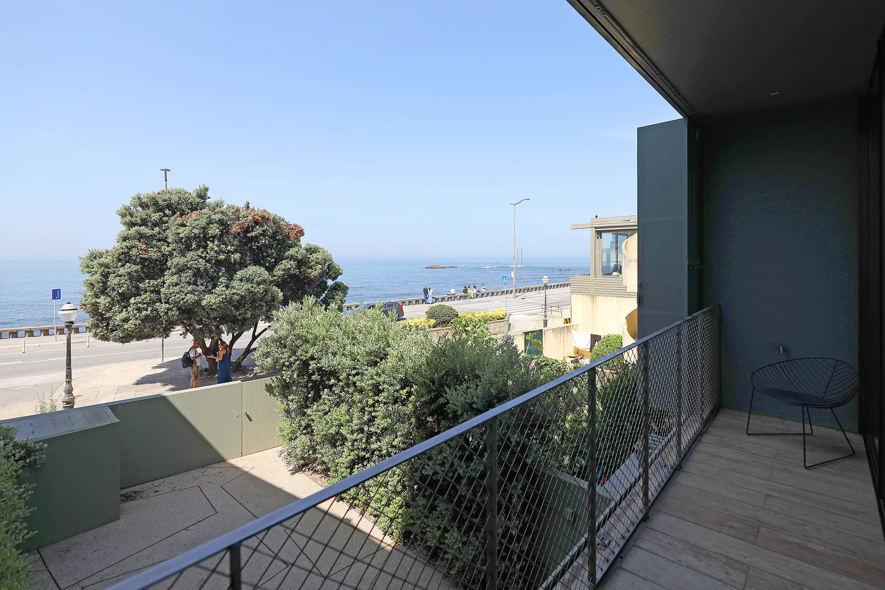 A balcony overlooks leafy trees, a coastal road, and the sea.