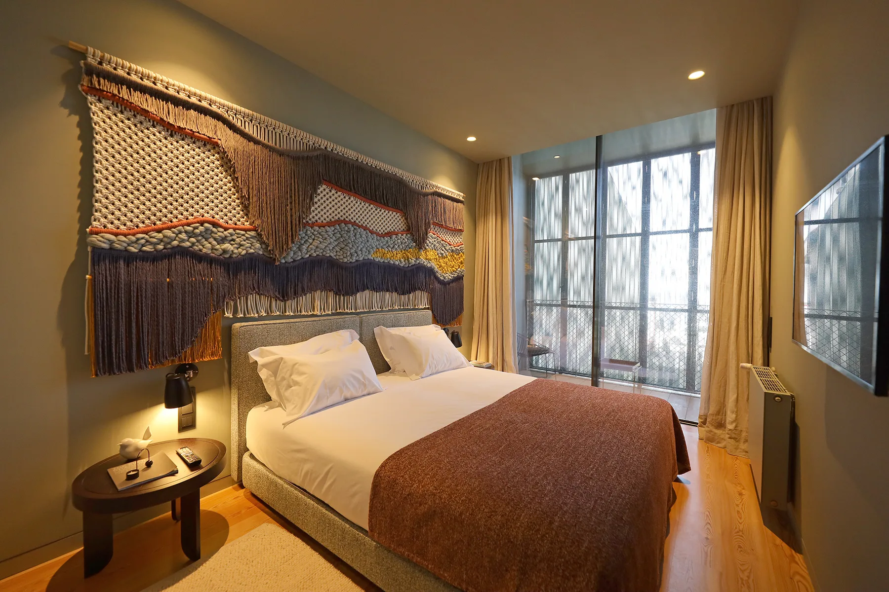 A bedroom with a bed, textured wall hanging, and tall curtained windows.