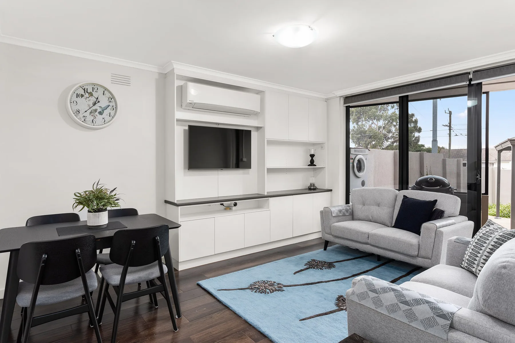 A bright living and dining area features a television wall and seating beside large windows.