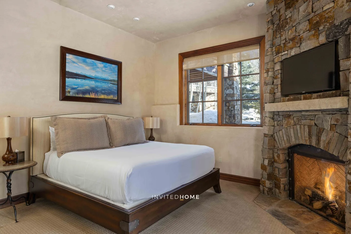 A bedroom with a bed beside a stone fireplace and a large window.