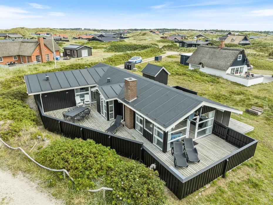 Una casa de techo oscuro con una amplia terraza de madera está entre dunas cubiertas de hierba.