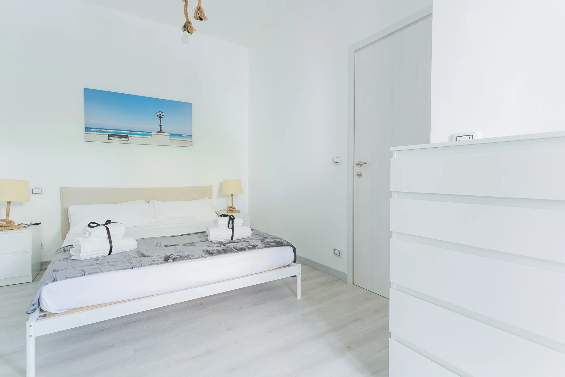 A bright bedroom with a double bed, bedside tables, and a coastal picture on the wall.