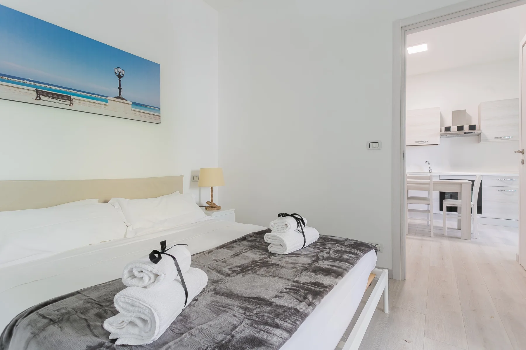 A bright bedroom with a neatly made bed and an open doorway to the kitchen.