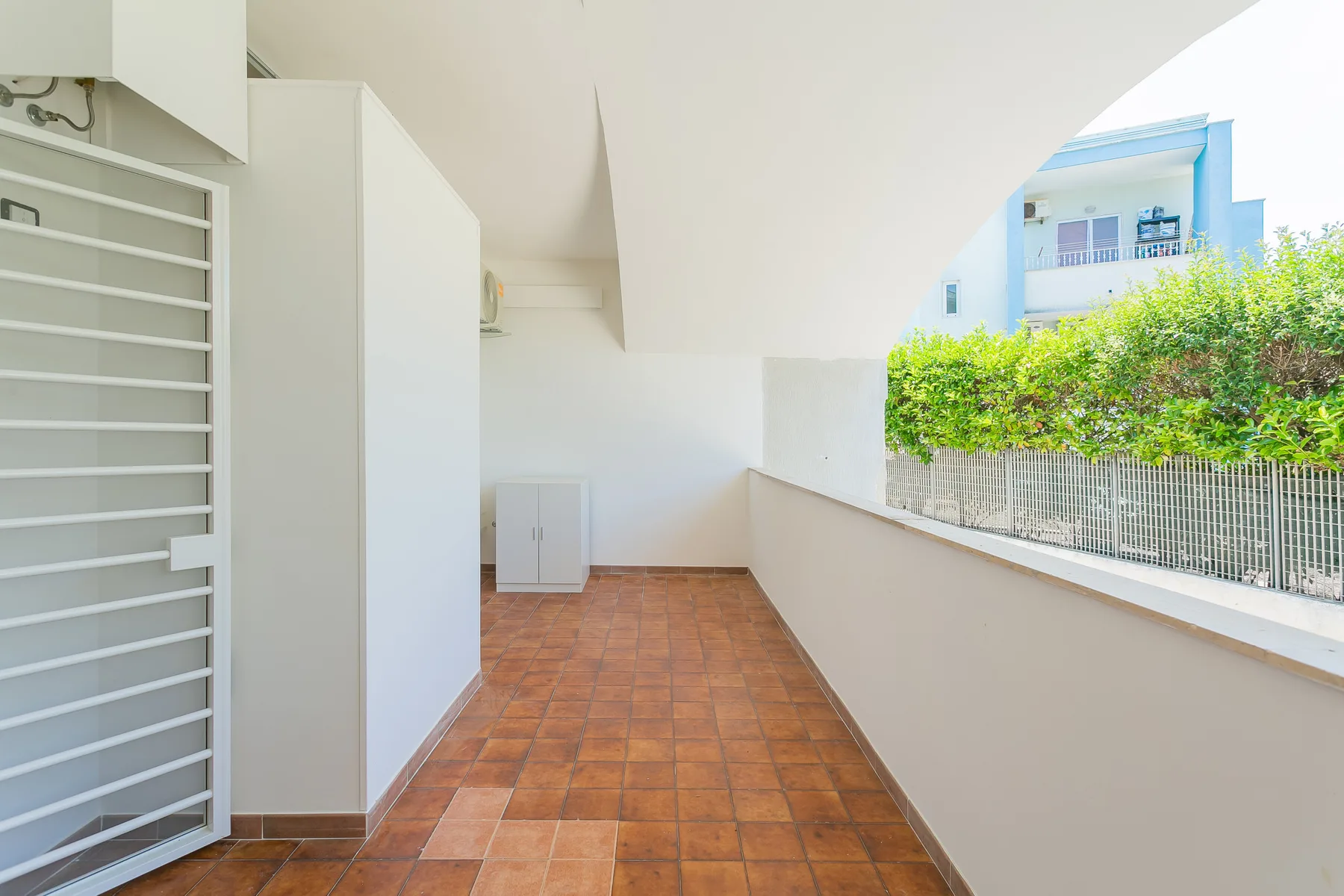 A covered outdoor walkway with terracotta-colored floor tiles and a railing.